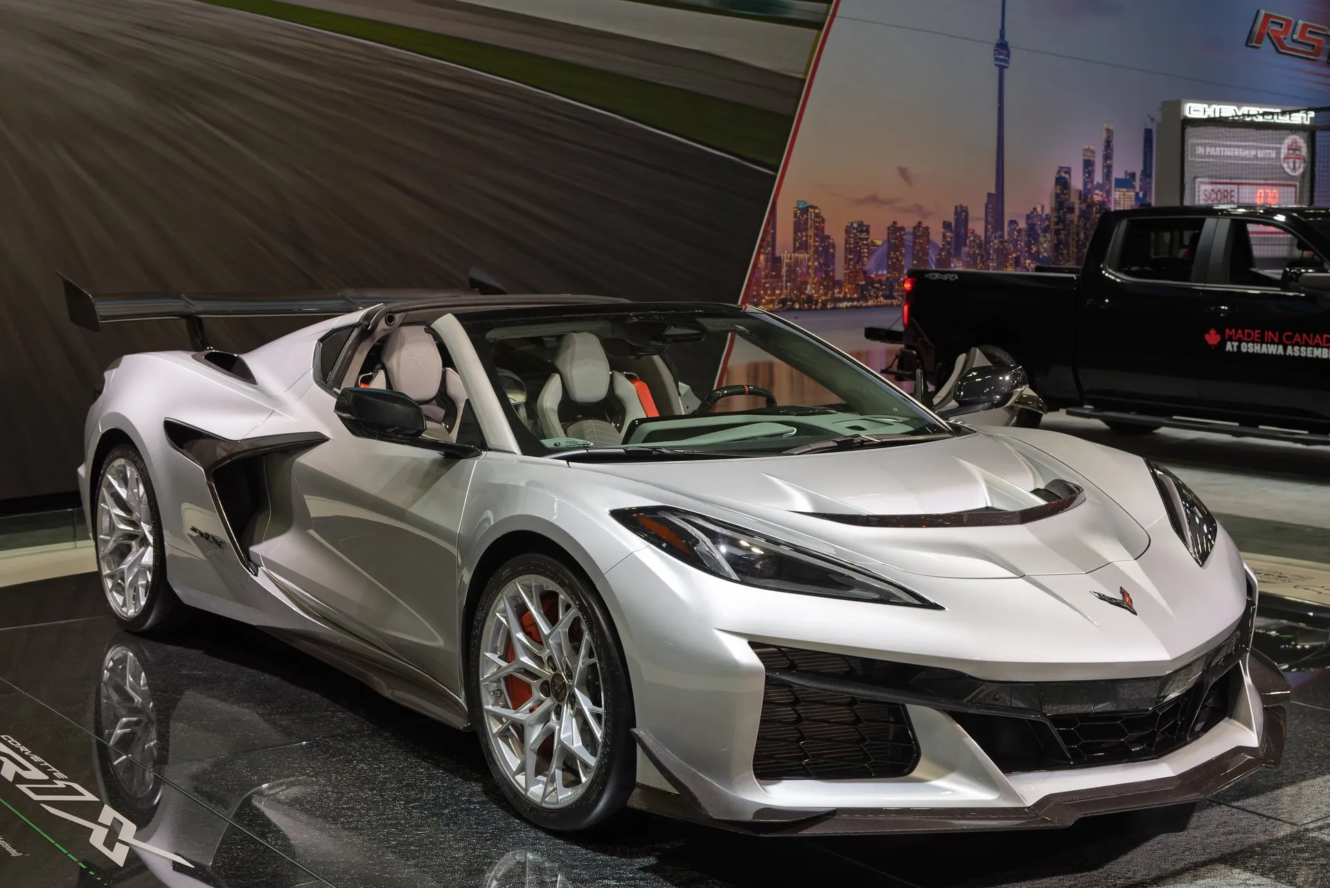 A silver C8 Corvette ZR1X on a glossy black auto show stand, seen from the front right: a black vent across the hood, silver wheels, red brake calipers, light gray seats and a tall rear wing, with ZR1X lettering on the floor and a black pickup truck and a city skyline backdrop behind it.