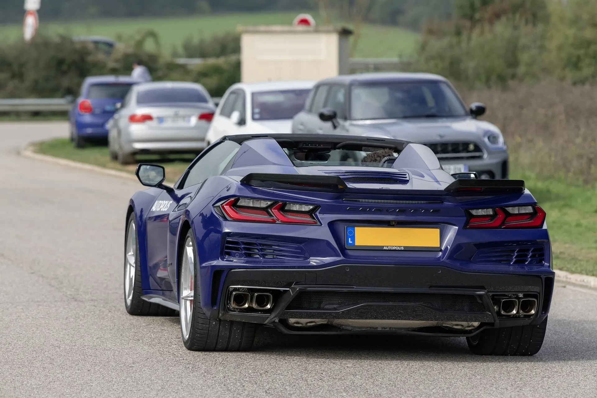 A dark blue C8 Corvette E-Ray convertible with its top down on a country road, seen from directly behind: wide rear fenders, a yellow European plate and four exhaust tips at the outer corners, with parked cars and hedges ahead of it.
