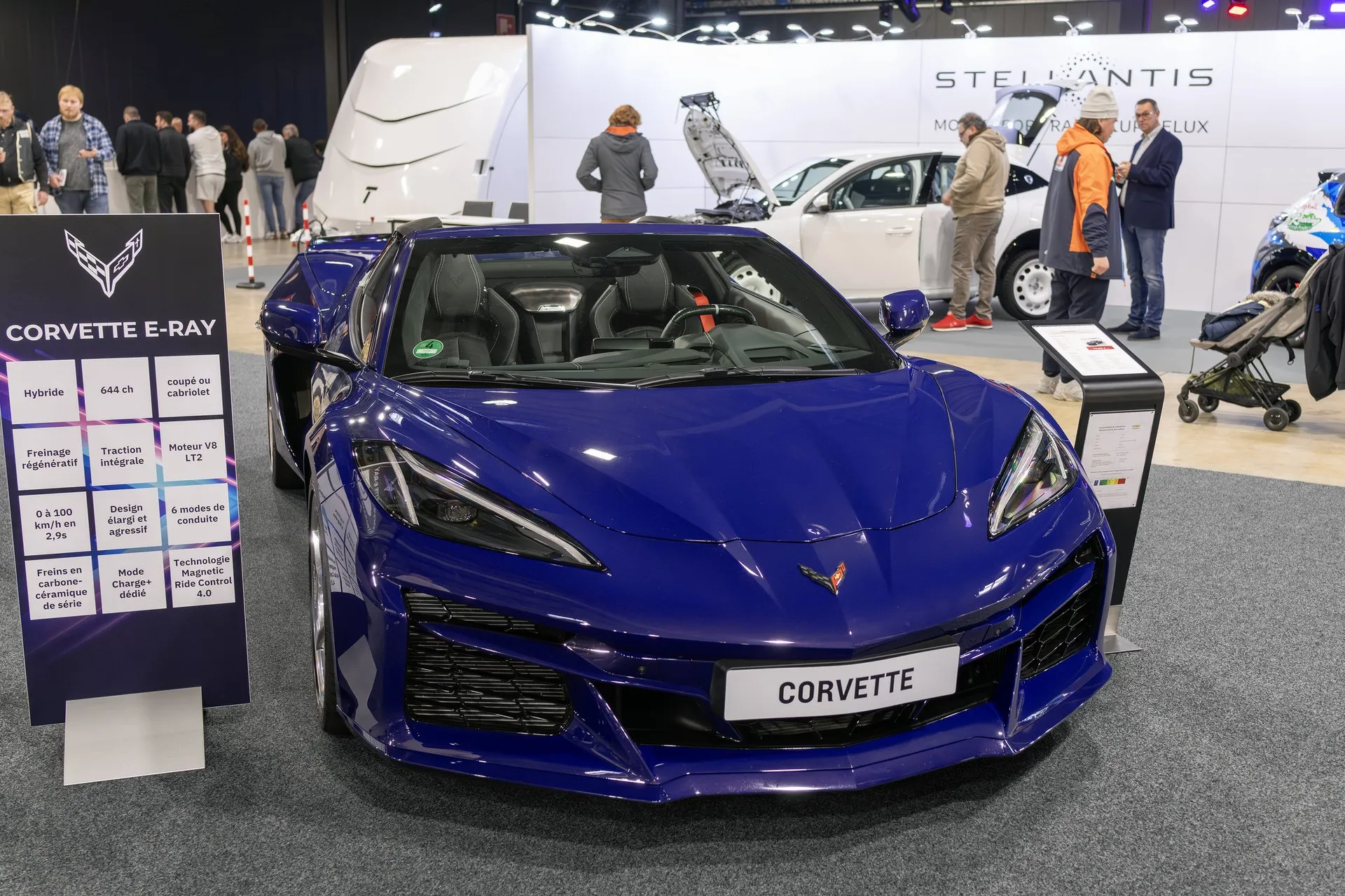 A dark blue C8 Corvette E-Ray seen head-on at an indoor show, with a Corvette name plate on its nose, black seats with red belts visible through the windshield, and a French-language display board titled Corvette E-Ray beside it.