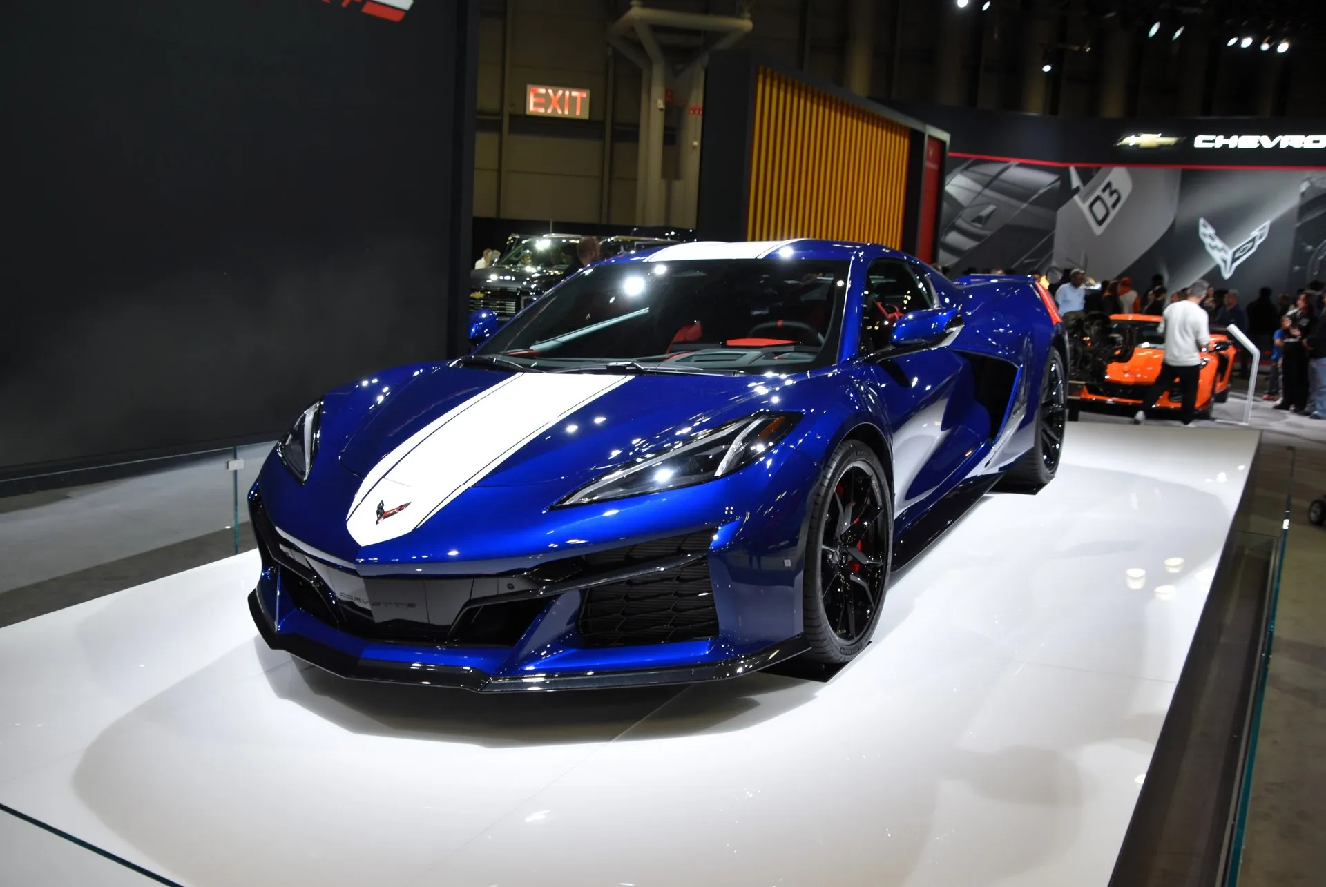 A blue C8 Corvette Grand Sport coupe with a white center stripe over the hood and roof, black wheels and red brake calipers, on a white display stand at an auto show, seen from the front left, with an orange Corvette and visitors behind it.