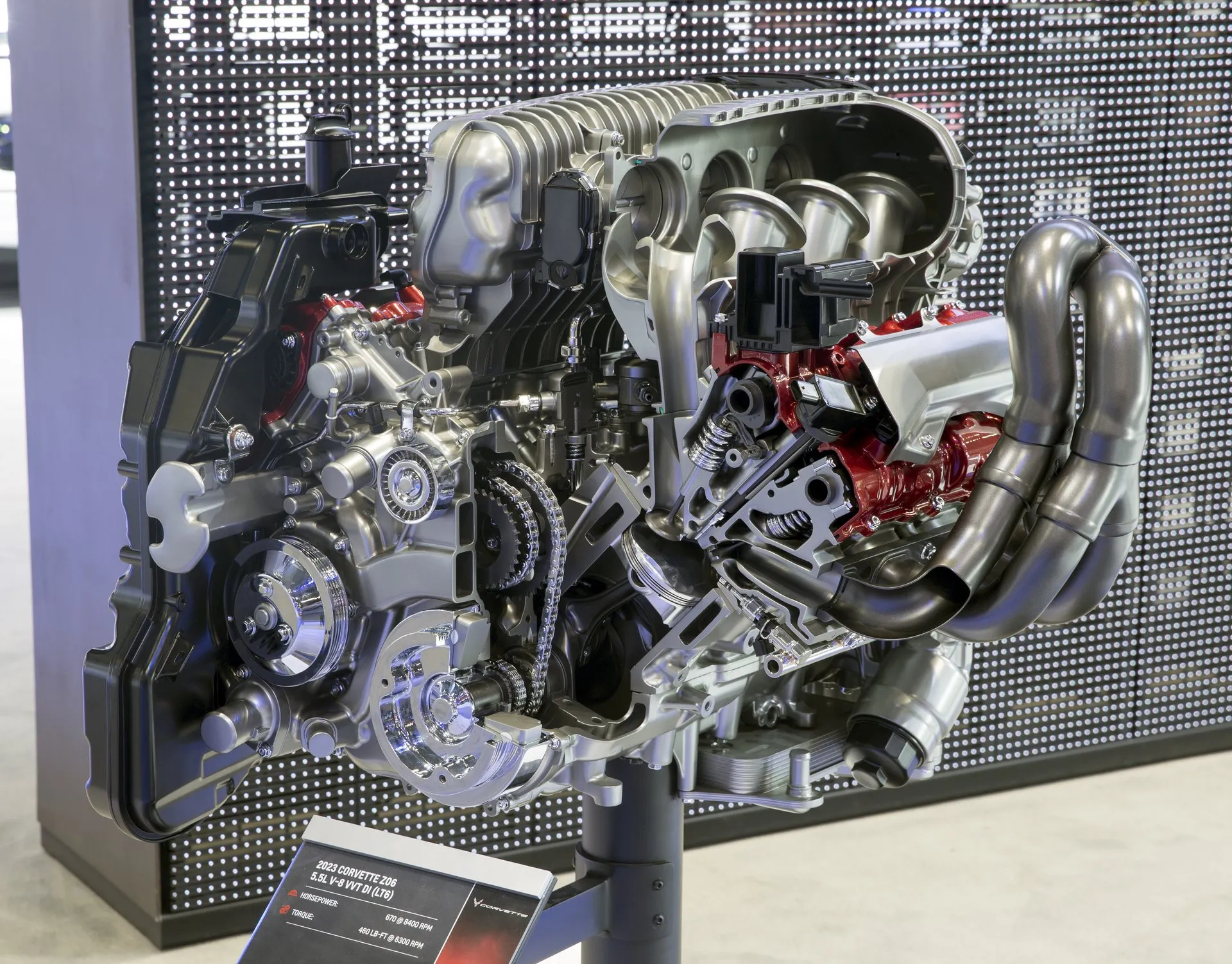 A cut-away V8 engine on a display stand in front of a perforated wall, opened up to show its intake runners, timing chains, valve springs, red-painted cylinder head parts and exhaust headers, with a placard at its base naming the 2023 Corvette Z06's 5.5-liter LT6.