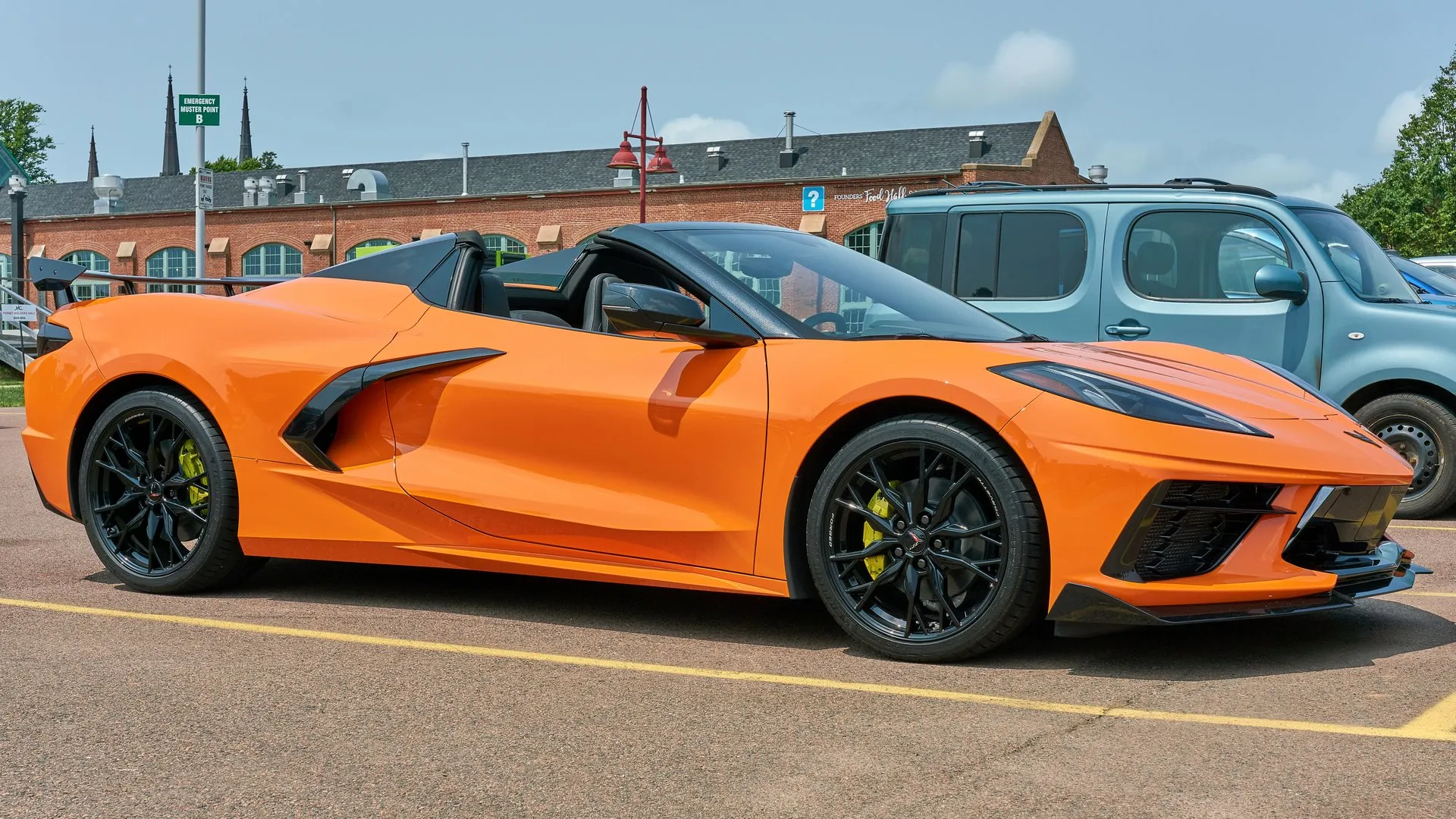 An orange C8 Corvette Stingray convertible with its hardtop folded away, black wheels, yellow brake calipers and a tall rear wing, parked in a lot beside a blue van, seen from the front right.