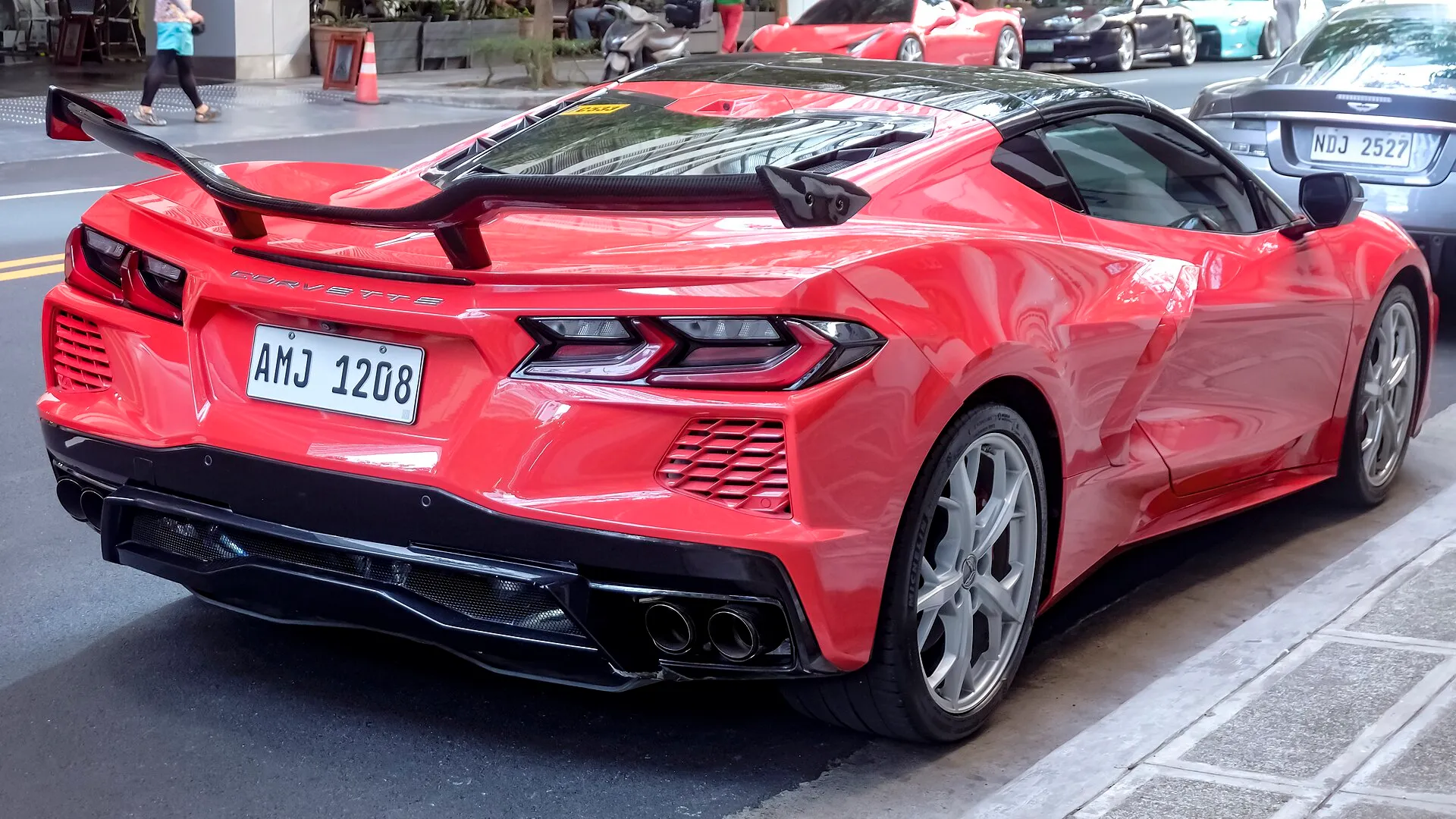Red C8 Corvette Stingray coupe seen from the rear right corner, parked at a curb, with a tall black rear wing standing on two uprights above the glass engine cover behind the cabin.