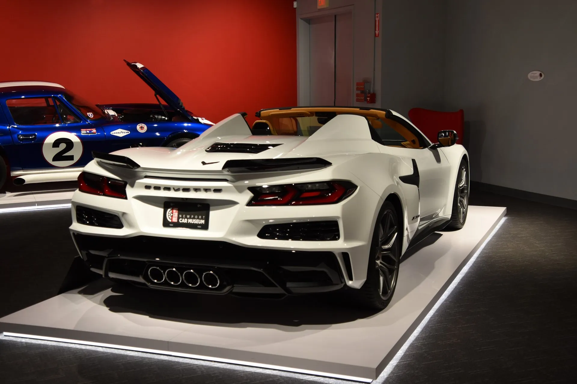A white C8 Corvette Z06 convertible with its top down and a tan interior on a lit display platform in a museum, seen from the rear right, with four exhaust tips in the middle of the bumper, beside a blue vintage Corvette race car numbered 2 with its hood up.