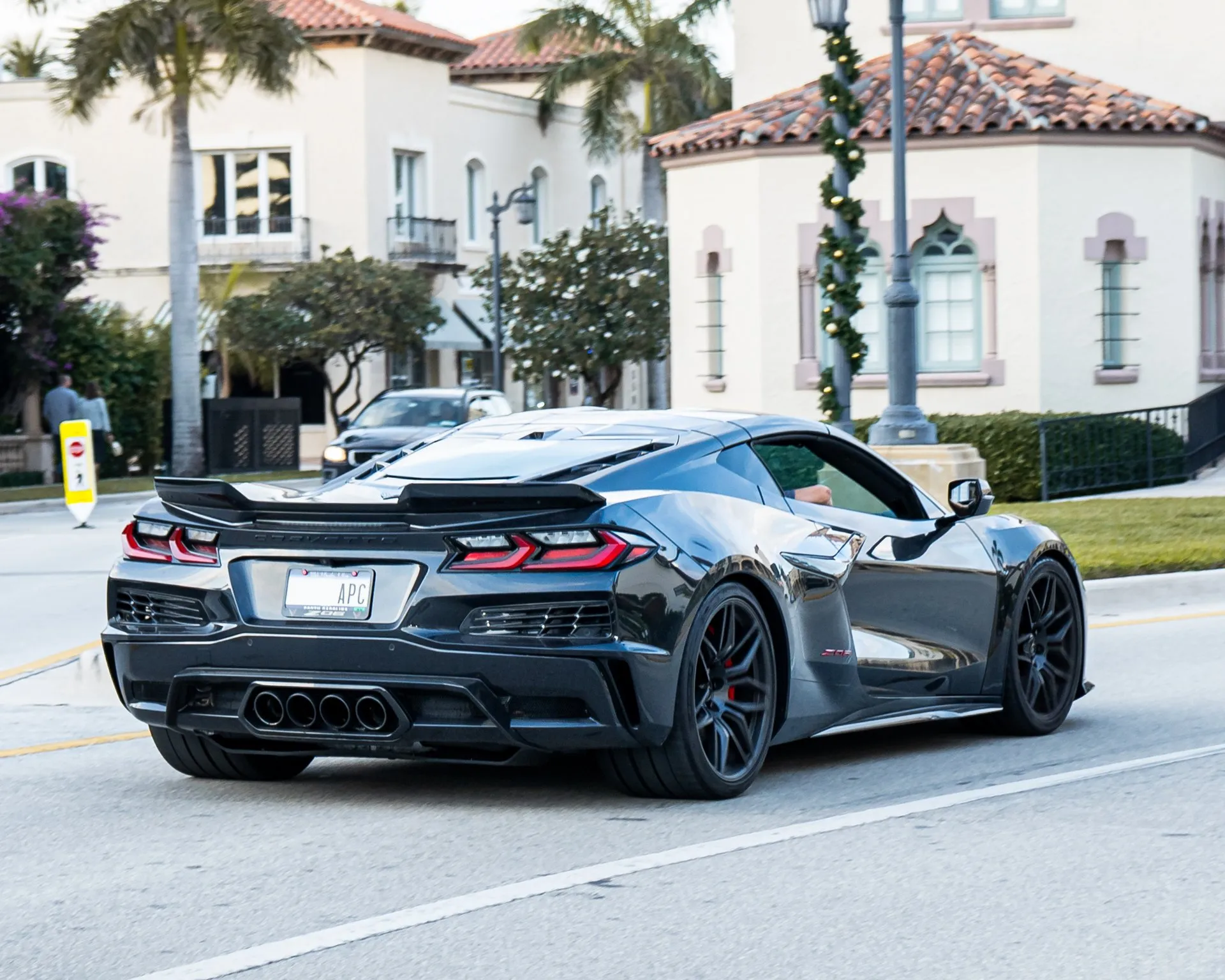 A dark gray C8 Corvette Z06 coupe driving away on a street lined with palm trees and stucco buildings, seen from the rear right, with four exhaust tips in the middle of the rear bumper, black wheels and red brake calipers.