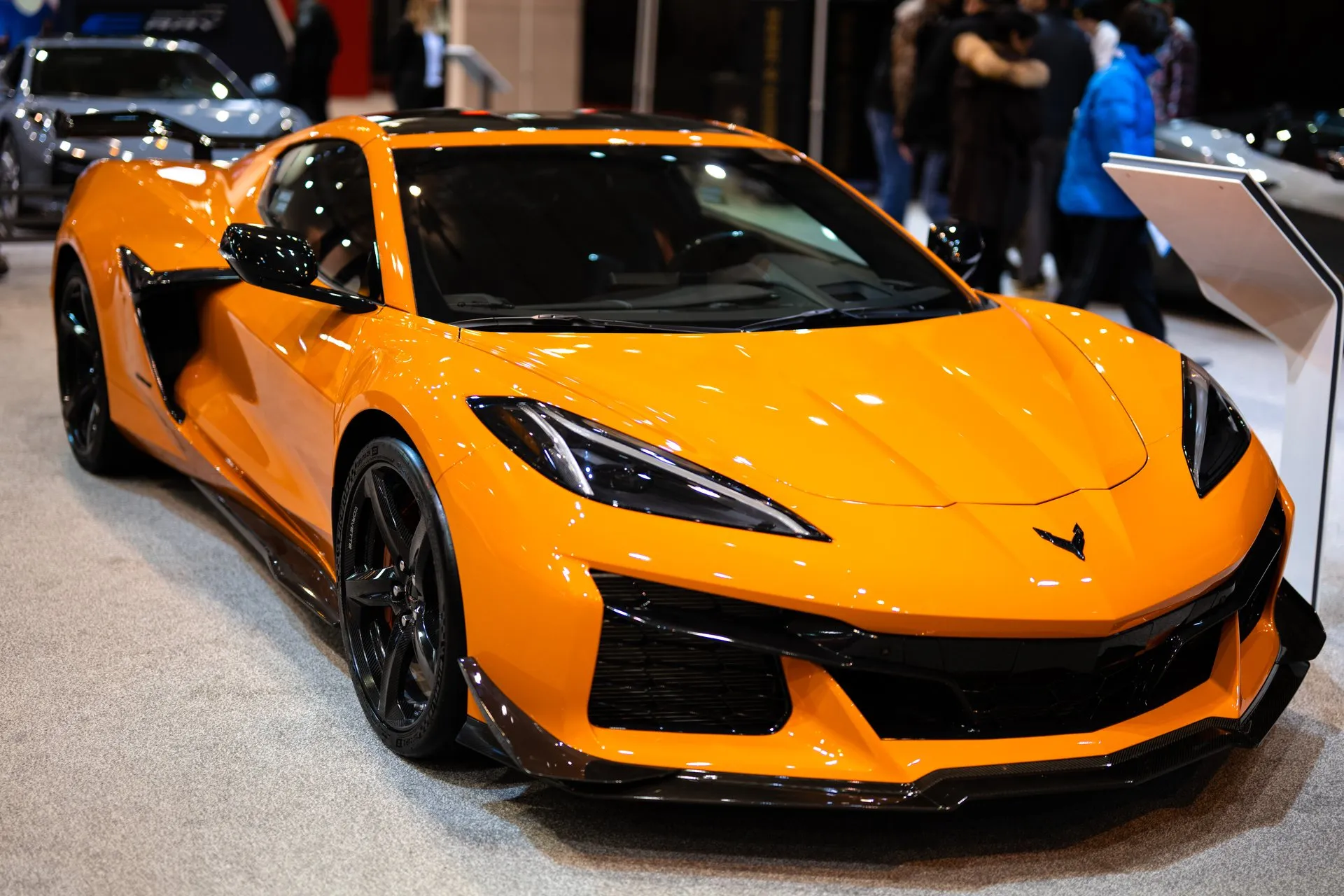 An orange C8 Corvette Z06 coupe with black wheels, a black carbon front splitter and small fins at the corners of the front bumper, on an auto show floor with visitors and a silver Corvette behind it, seen from the front right.